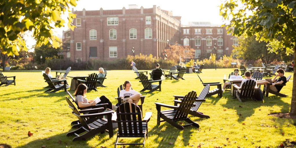 Students are sitting outside, with Harkins Hall in the background, in Adirondack chairs.